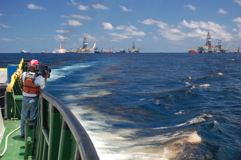 A man on a boat with a camera pointed toward several vessels and structures in the water.