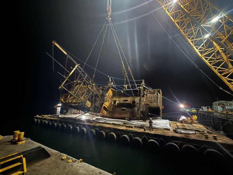 A fishing vessel lifted by a crane being rested atop a large barge at night.