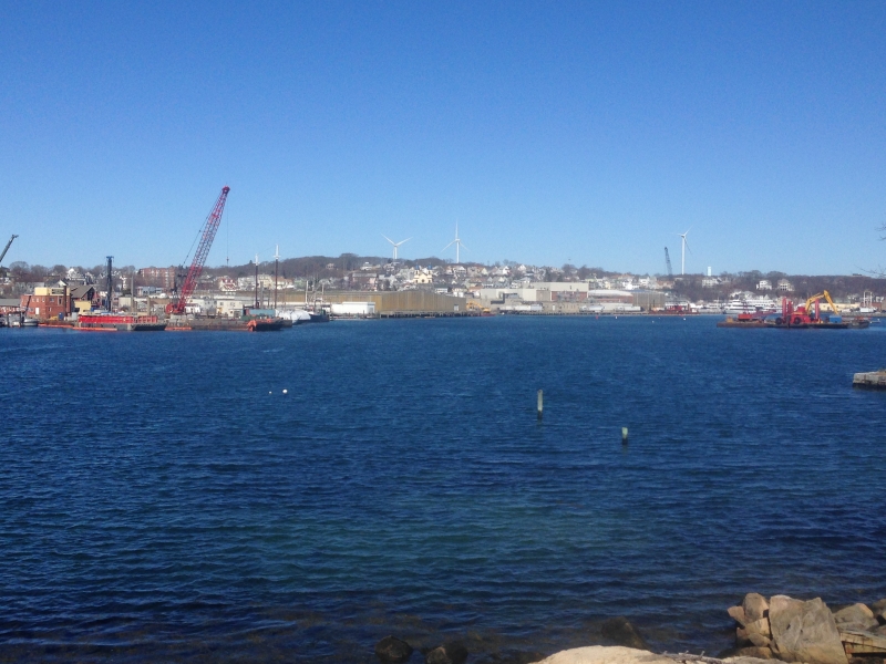 Gloucester harbor with deep blue waters in the foreground, featuring construction cranes, docks, and industrial buildings along the shoreline.
