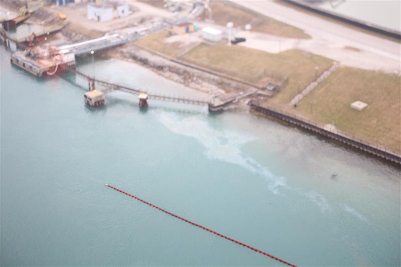 Aerial image of spill boom (oil containment tool) floating at the surface of water along a waterway with an industrial facility close by.