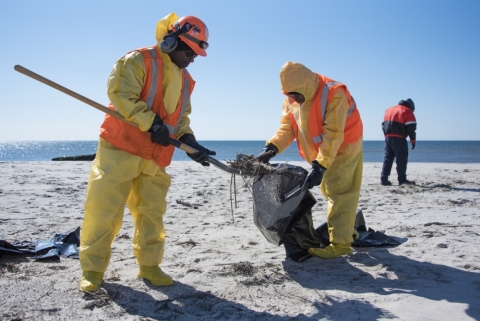 Two people in orange and yellow response gear picking up a black substance with a shovel. 