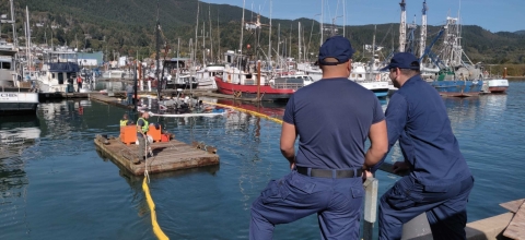 Two people oversee pollution boom placement at a marina where a sunken boat is visible above the water.