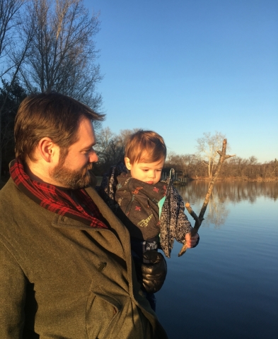 A man holding a baby with a body of water in the background. 
