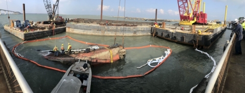 Several people on an overturned vessel surrounded by pollution boom.