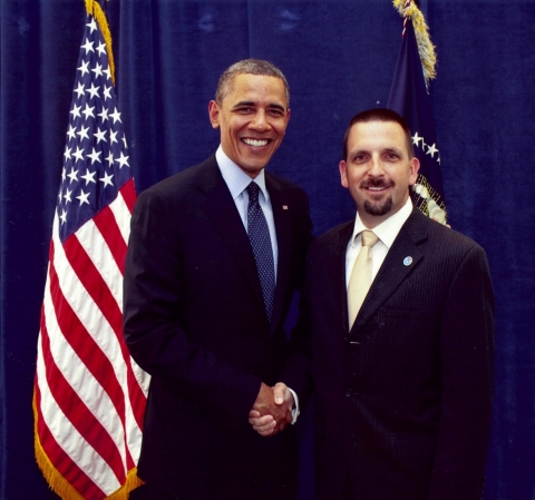 Two men shaking hands with an American flag behind them.