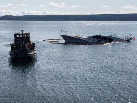 A small boat next to a partially sunken vessel surrounded by pollution boom.