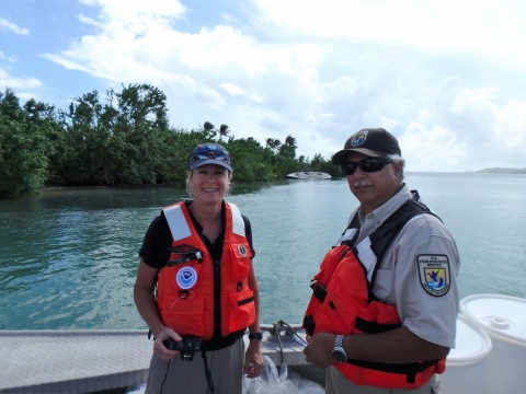 A man and a woman in life jackets on a boat with a derelict vessel along a green shoreline in the background.