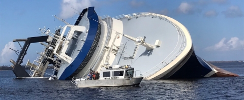 A large vessel on its side in the water with a smaller boat in front of it.