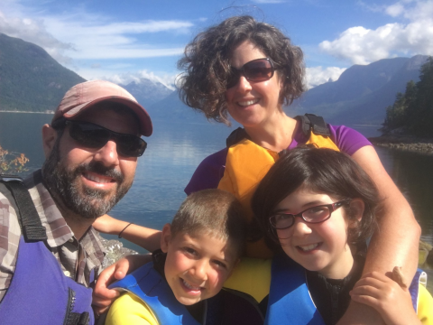 A man, a woman, and two kids all wearing life vests with a body of water and a mountain landscape in the background.