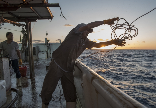 A silhouette of a man throwing a rope on a boat out at sea. 