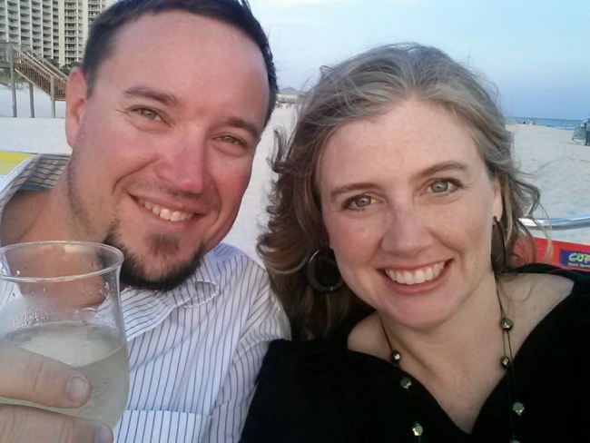 A man and a woman posing for a photo with a beach behind them. 