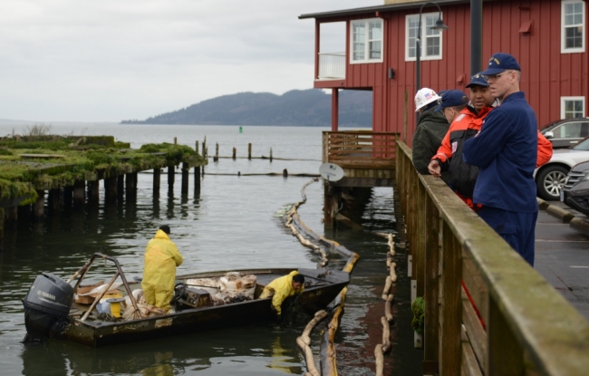 Coast Guard members standing on a dock looking out as responders place boom around the pier from a dinghy. 