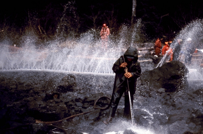 Cleanup workers and hoses spraying out water working to clean off black rocks on a shoreline.