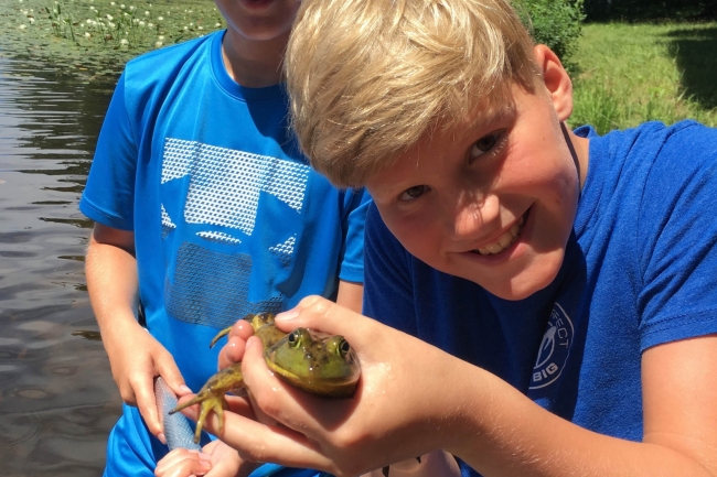 A young boy standing next to another boy in a body of water holding a frog.