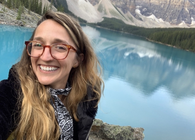 A woman with a body of water and mountains behind her.