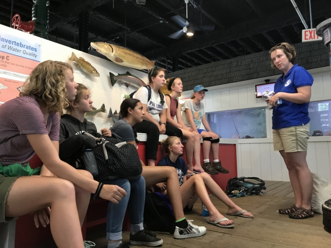 A woman standing on the right talking to a group of kids on the left.