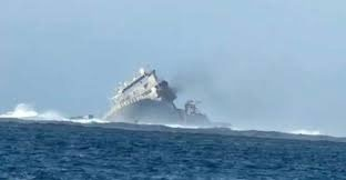 A vessel in the ocean tilts on its side. Smoke is coming out of the vessel and it is surrounded by waves.