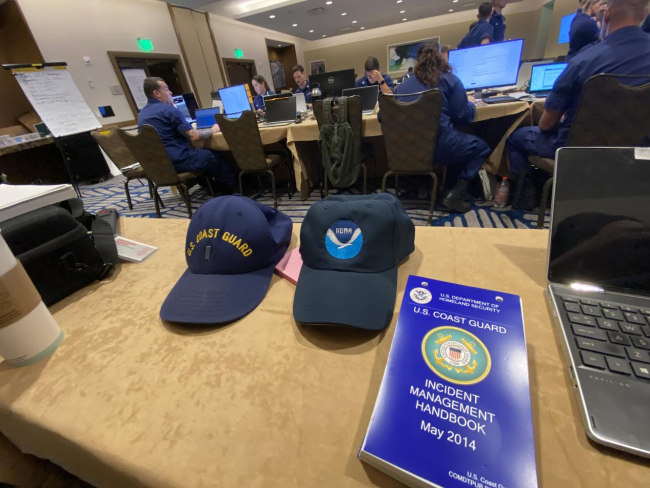 Two hats, one branded with the U.S. Coast Guard and one branded with NOAA's logo, sit beside each other on a table next to an incident management handbook. The table is within an area command post responding to a hurricane.