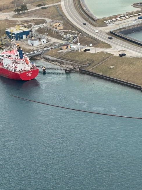 Aerial image of spill boom (oil containment tool) floating at the surface of water behind a docked vessel docked along a waterway with an industrial facility close by.