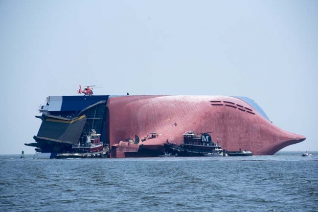 A large vessel on overturned on its side with boats around it and a helicopter on top. 