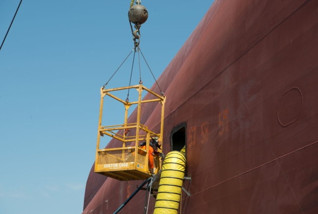 A man in a crane cage reaching out toward a large duct hose coming out of a vessel. 
