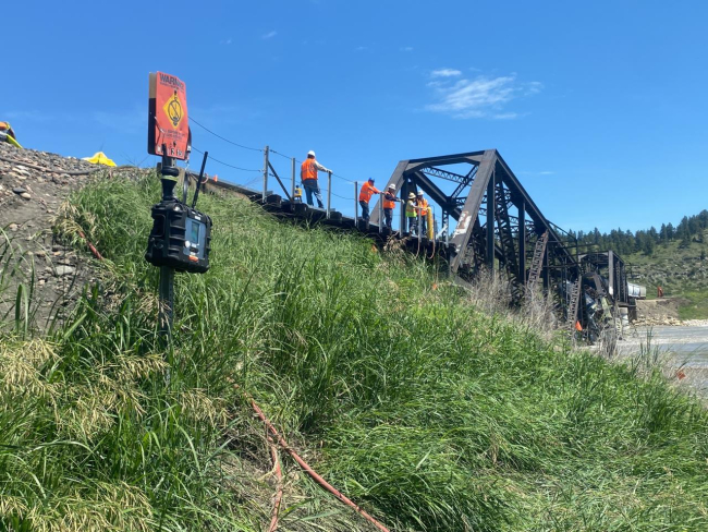 Grassy bank along railroad tracks with workers on a bridge.