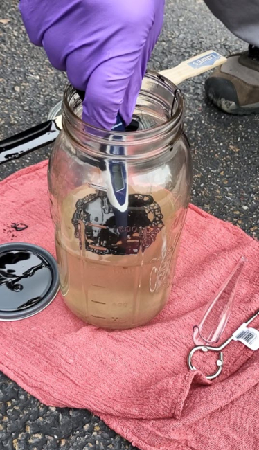 Gloved hand scooping oil sample from a jar.