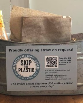 A metal tin sits on a wooden table and holds hot sauce and napkins. There is a sign on the tin that reads “Proudly offering straw on request! Skip the plastic.”