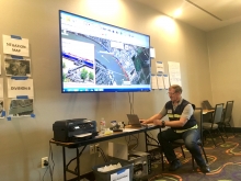 A man using a laptop at a table beneath a large screen depicting a map. 