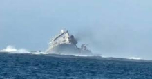 A vessel in the ocean tilts on its side. Smoke is coming out of the vessel and it is surrounded by waves.