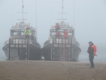 A man stands on a sandy beach with four crew members behind him in two boats searching the area.