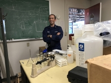 A man standing in a lab with a chalkboard behind him. 