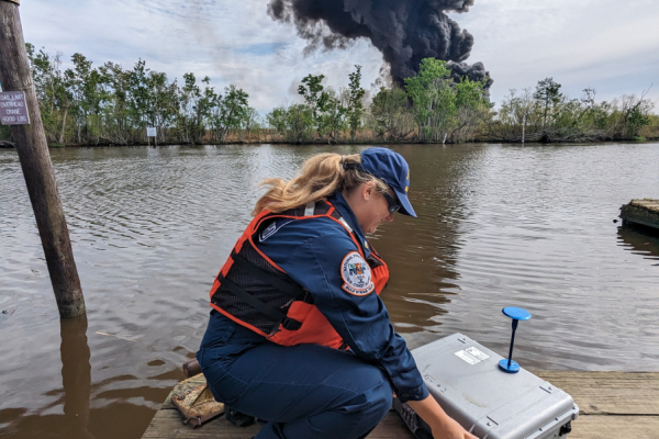 A woman on a dock setting up monitoring equipment with a black smoke stack from burning oil in the distance.